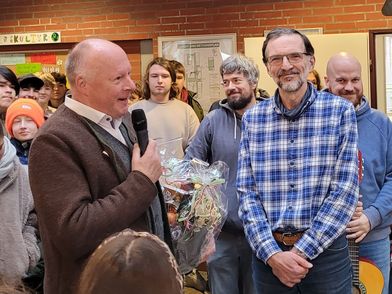 In der Schulaula mit roter Backsteinwand und einer Uhr mit Mosaikrahmen an der Wand steht Bürgermeister Dr. Jens Bülthuis mit Mikrofon und einem Geschenk in der Hand. Daneben steht Klaus Böger in blau-kariertem Hemd. Um sie herum versammeln sich mehrere Personen, einige mit Instrumenten wie einer Gitarre.