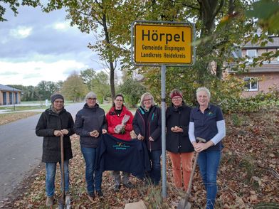 Sechs Frauen stehen an einem Ortsschild von Hörpel, Gemeinde Bispingen, Landkreis Heidekreis, eine hält eine Schaufel, andere tragen eine dunkelblaue Decke mit Aufdruck 'LandFrauen Bispingen'.