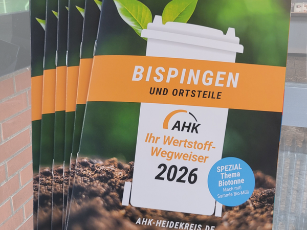 Mehrere gedruckte Broschüren liegen übereinander. Auf dem Titel ist ein weißer Becher mit Deckel abgebildet, aus dem zwei grüne Blätter wachsen. Der Hintergrund zeigt unscharf Erde und grünes Laub. Auf einem orangefarbenen Balken steht in Großbuchstaben 'BISPINGEN UND ORTSTEILE'. Darunter befindet sich das Logo 'AHK' und der Text 'Ihr Wertstoff-Wegweiser 2026'. Ein blauer Kreis mit weißer Schrift weist auf das Spezialthema 'Biotonne' hin mit dem Hinweis 'Mach mit! Sammle Bio-Müll'. Unten steht die Webadresse 'AHK-HEIDEKREIS.DE'.