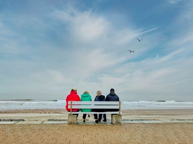 Vier Personen von hinten. Sie sitzen mit bunten Winterjacken auf einer Parkbank an der Standpromenade Norderney. 