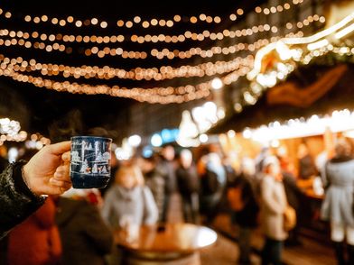 Hand, die eine Tasse Glühwein hält im Fokus und das beleuchtete Einbecker Weihnachtsdorf dahinter.