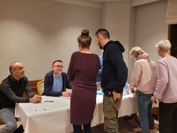 Samer Tannous und Gerd Hachmöller sitzen an einem Tisch mit weißer Tischdecke, vor ihnen stehen mehrere Personen in einem Raum mit Fenster und Pflanze.