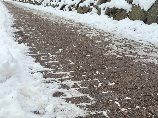 Ein gepflasterter Weg mit einer dünnen Schneeschicht ist zu sehen. Der Weg verläuft schräg im Bild und ist an einer Seite von einer niedrigen Steinmauer mit aufliegendem Schnee begrenzt. Auf dem Weg sind vereinzelt Schneereste verteilt, während die Pflastersteine größtenteils freigeräumt sind. Am Bildrand liegt eine dickere Schneeschicht neben dem Weg.