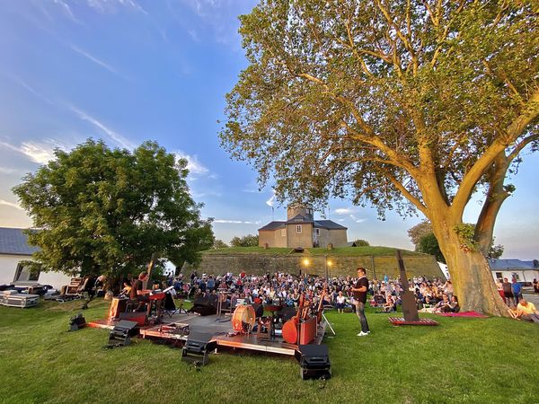 Open-Air-Konzert auf einer grünen Wiese vor einer historischen Festung bei Sonnenuntergang.