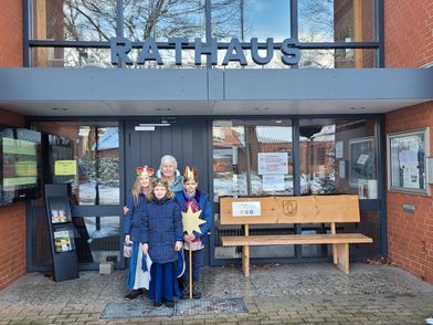 Vor dem Eingang des Bispinger Rathauses stehen mehrere als Sternsinger verkleidete Kinder. Die Kinder tragen bunte Umhänge und Kronen, eine Person hält einen goldenen Stern auf einem Stab. Im Hintergrund sind Glastüren mit Aushängen, ein Holzbank und ein Schaukasten zu sehen. Über dem Eingang steht in großen Buchstaben das Wort 'RATHAUS'. Auf dem Boden liegt etwas Schnee.