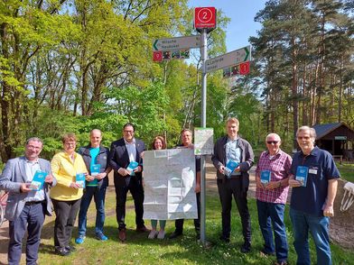 Vorstellung der neuen Südheide-Gifhorn-Radwanderkarte Personengruppe mit einer aufgeklappten Radkarte vor einem Fahrradwegweiser mit Knotenpunkt in der Natur.