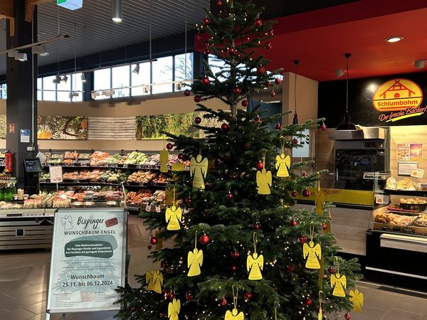 Weihnachtsbaum mit roten Kugeln und gelben Engelanhängern in einem Supermarkt, daneben ein Schild mit Informationen zur Wunschbaum-Engel Aktion.
