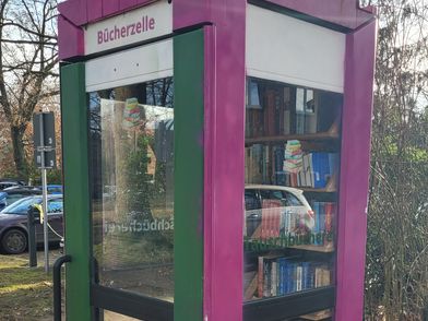 Eine kleine, freistehende Bücherzelle in Form einer Telefonzelle mit gläsernen Wänden und einer grünen Tür. Das Dach und die Ecken sind in einem dunklen Pink gehalten. Im Inneren sind mehrere Regale mit Büchern gefüllt. Über der Tür befindet sich ein Schild mit der Aufschrift 'Bücherzelle'. Die Bücherzelle steht auf einem gepflasterten Untergrund, im Hintergrund sind Bäume ohne Blätter und ein geparkter blauer Pkw zu sehen.