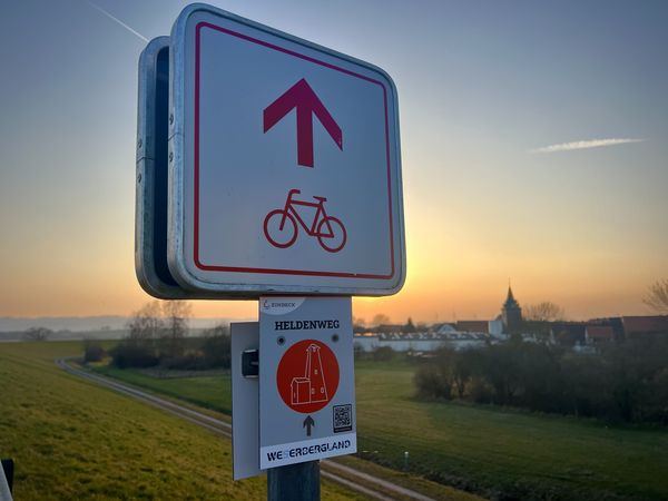 Wegschild zum Heldenweg vor einem Dorf mit Kirchturm bei Sonnenuntergang.