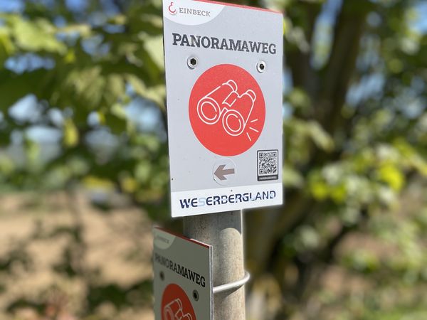 Weißes Schild für den Panoramaweg mit rotem Fernglas-Symbol und Richtungspfeil im Grünen.